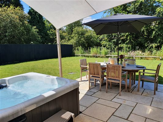 the patio in summer, hot tub to the left with awning above anbd wooden dining table and chairs with large black umbrella